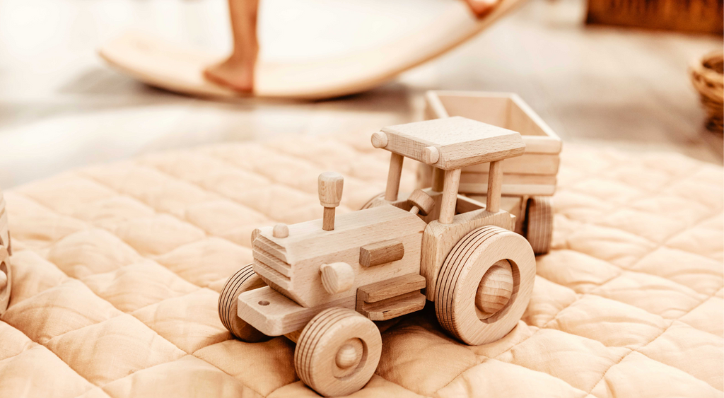Natural wooden toy tractor with trailer sitting on apricot coloured play mat.  Legs of young boy balancing on wooden wobble board shown in background.