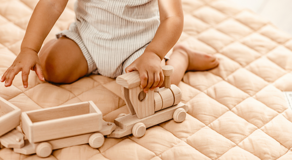 Infant kneeling on apricot coloured mat holding natural wooden toy train engine towing two carriages