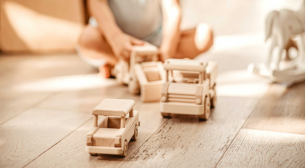 Boy sitting crossed legged on floor holding natural wooden toy bulldozer.  Natural wooden toy truck and car are shown in front.